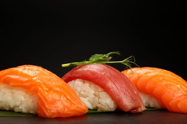 Close up three nigiri sushi with salmon and tuna fish served on fresh green banana leaf over black background, low angle side view